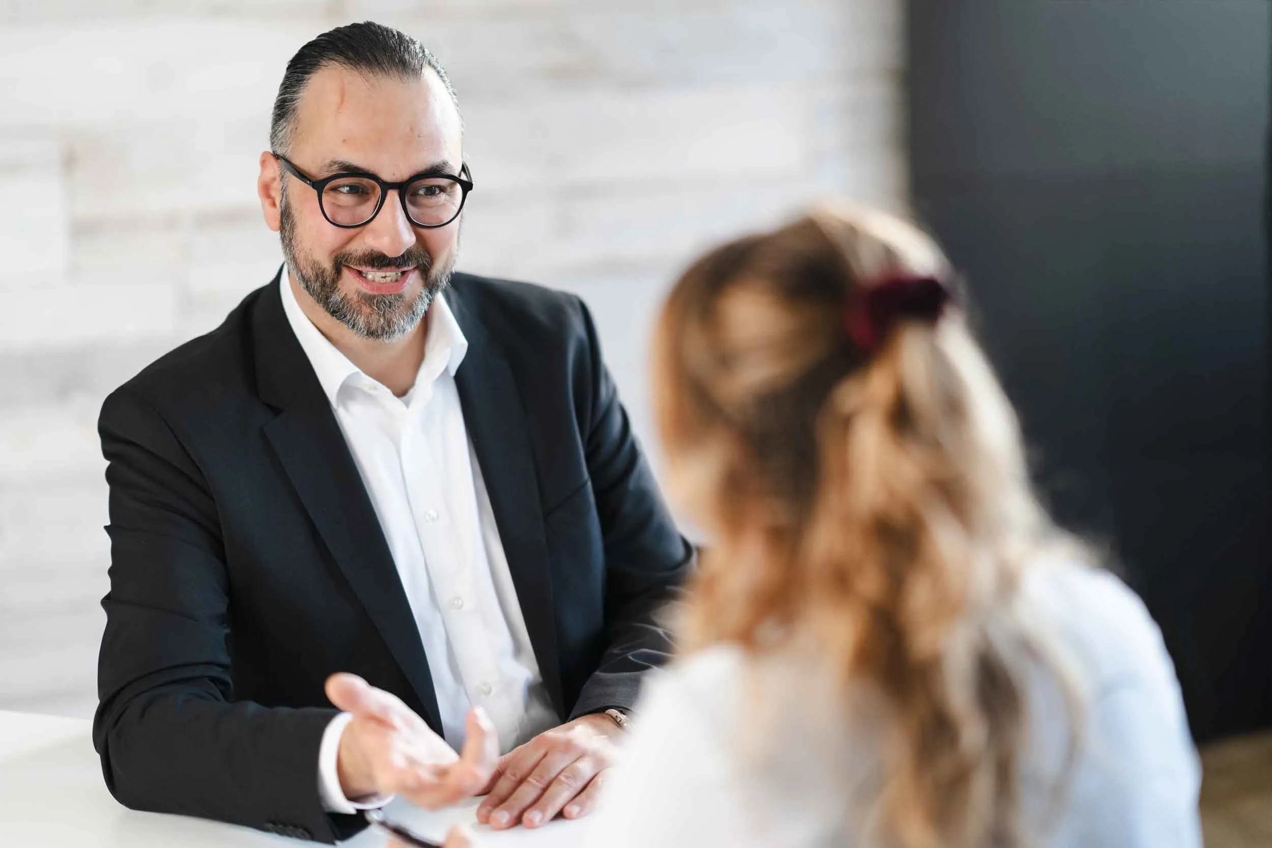 Ömer Özbekler, lächelnd, in schwarzem Anzug und weißem Hemd, hat Unterlagen vor sich liegen und berät Klientin